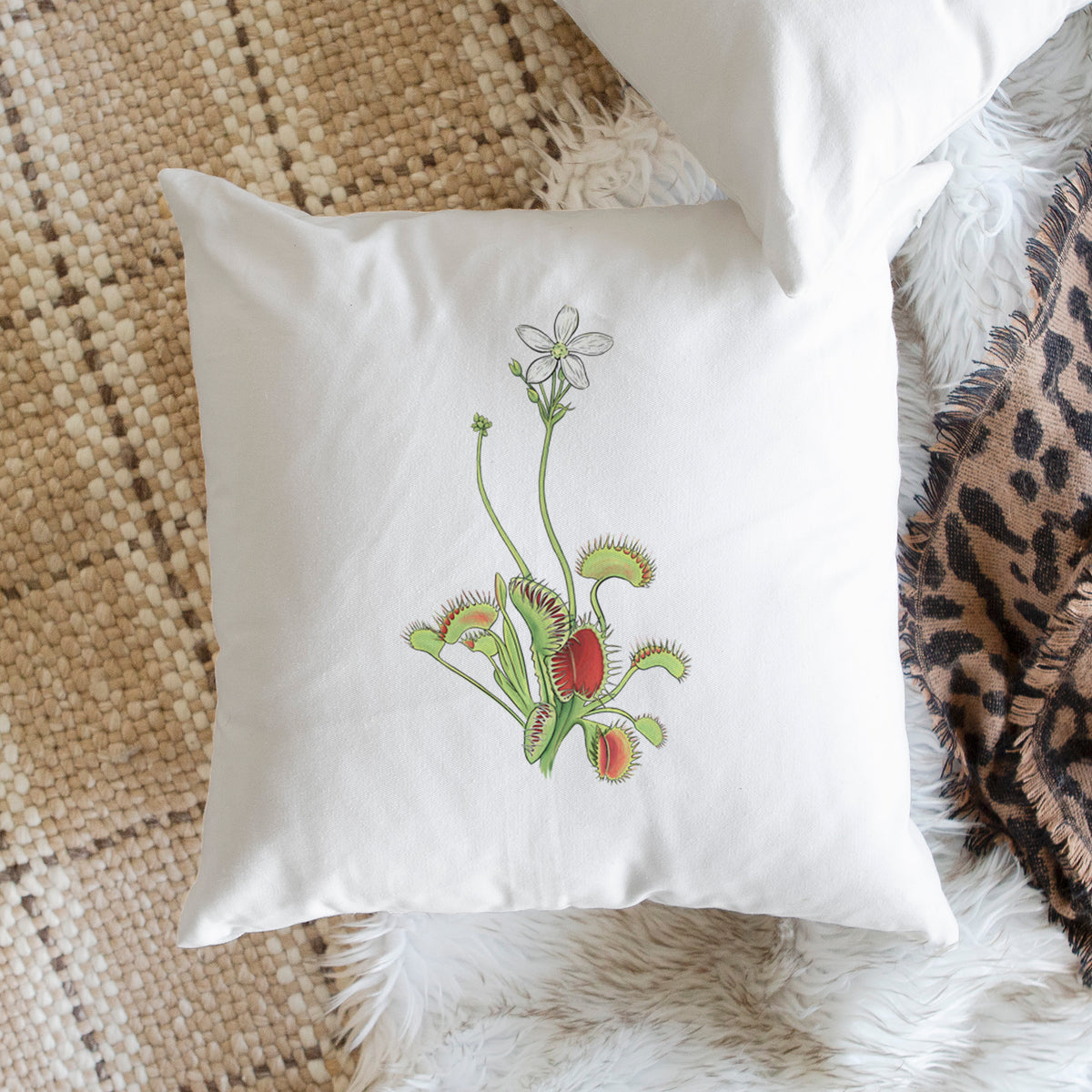 White pillow cover printed with a full-color illustration of a Venus flytrap plant with red traps and a small white flower, on a woven chair.
