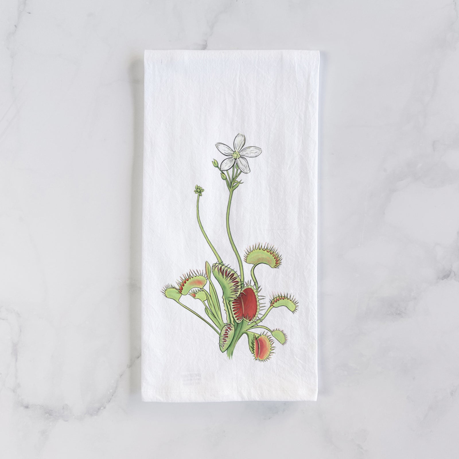 White cotton tea towel printed with a full-color illustration of a Venus flytrap plant with red traps and a small white flower, on a marble counter beside a round wooden cutting board and greenery.