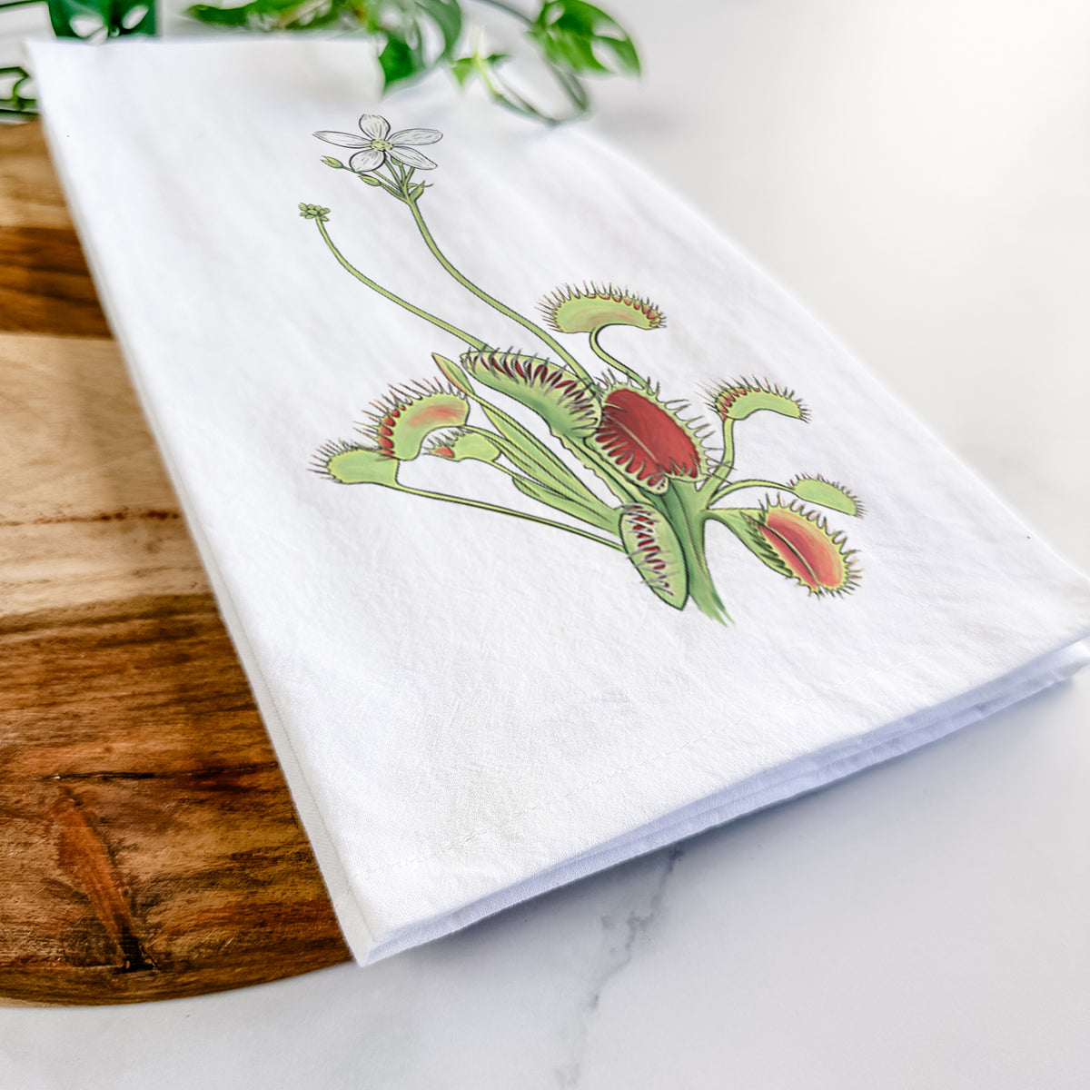 Close-up of a folded white tea towel on a wooden board, showing a full-color illustration of a Venus flytrap plant with red traps and a small white flower.