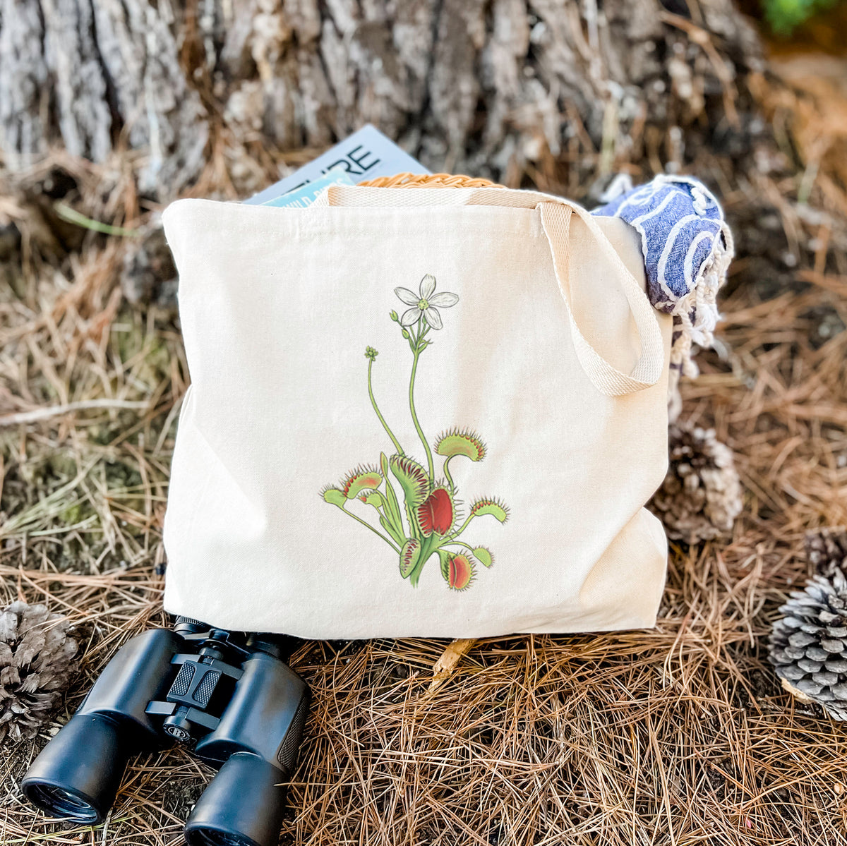 Canvas tote bag printed with a full-color illustration of a Venus flytrap plant with red traps and a small white flower, resting on dry grass beside a pair of binoculars.
