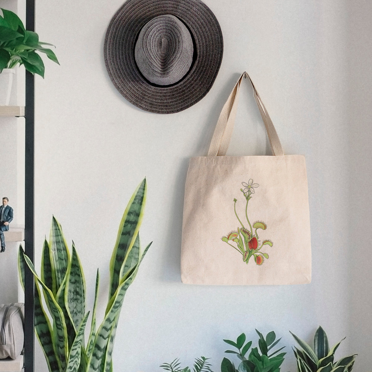 Canvas tote bag printed with a full-color illustration of a Venus flytrap plant with red traps and a small white flower, hanging on a wall hook below a hat, next to potted plants.