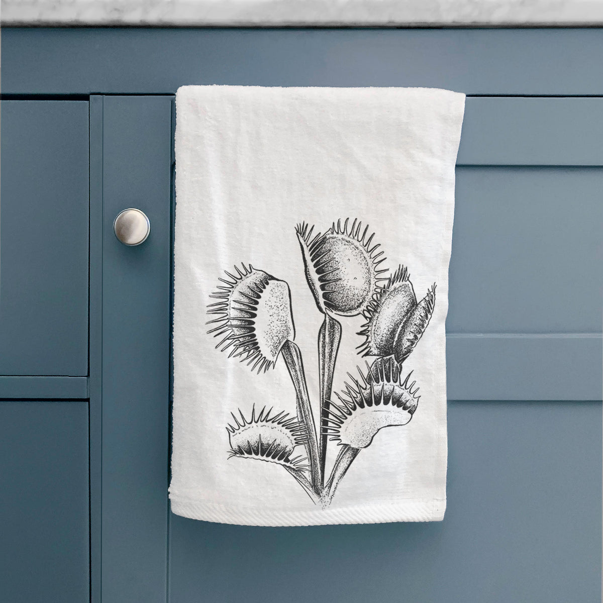 White hand towel printed with a black ink illustration of a Venus flytrap plant with open traps, hanging on a cabinet door.