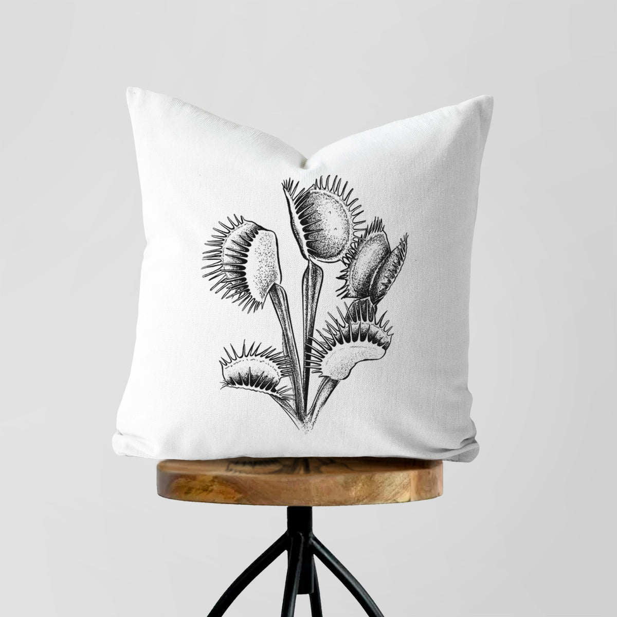 White pillow cover printed with a black ink illustration of a Venus flytrap plant with open traps, on a wooden stool.