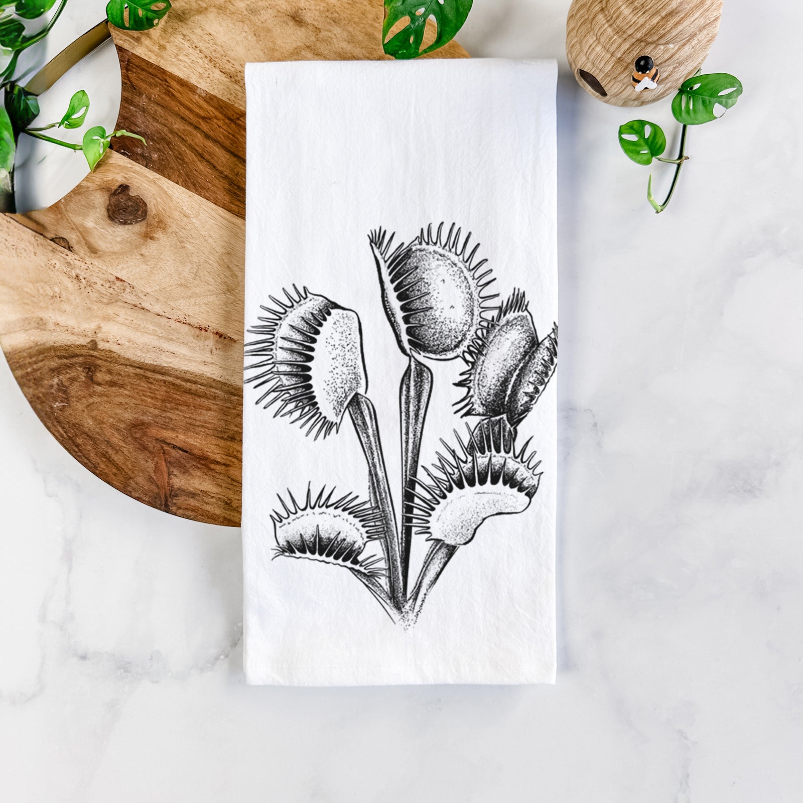 White cotton tea towel printed with a black ink illustration of a Venus flytrap plant with open traps, on a marble counter beside a round wooden cutting board and greenery.
