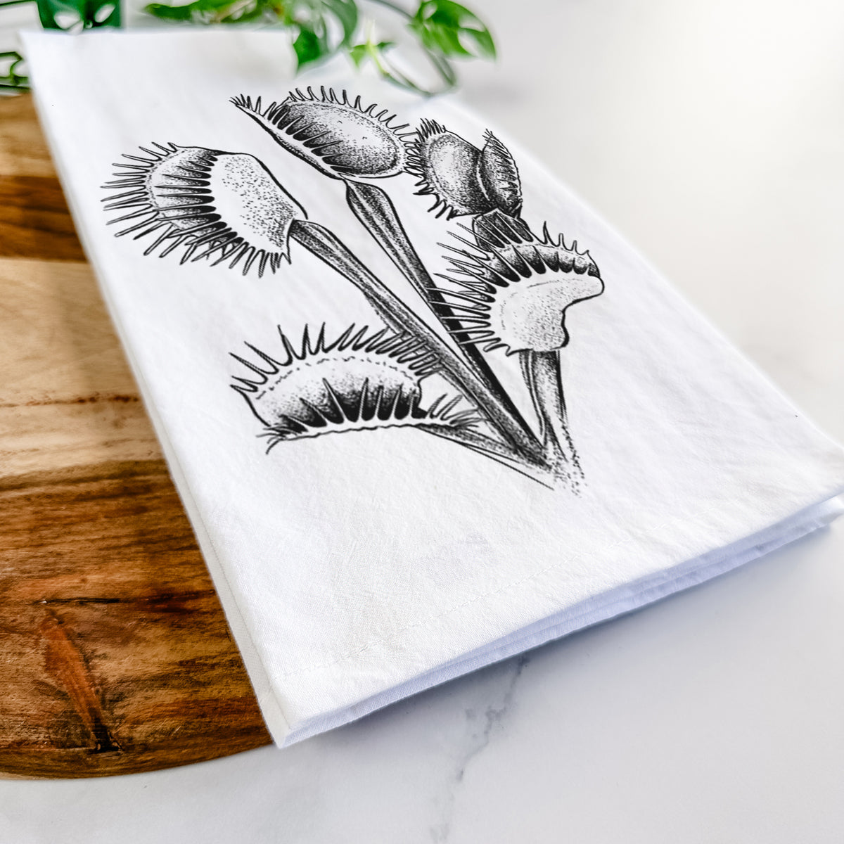 Close-up of a folded white tea towel on a wooden board, showing a black ink illustration of a Venus flytrap plant with open traps.