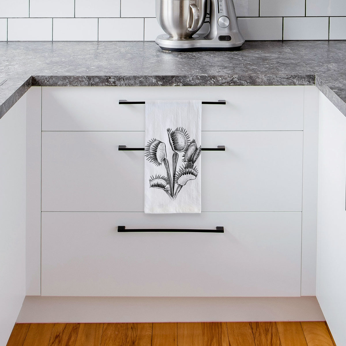 White tea towel printed with a black ink illustration of a Venus flytrap plant with open traps, hanging from a drawer handle in a bright white kitchen.