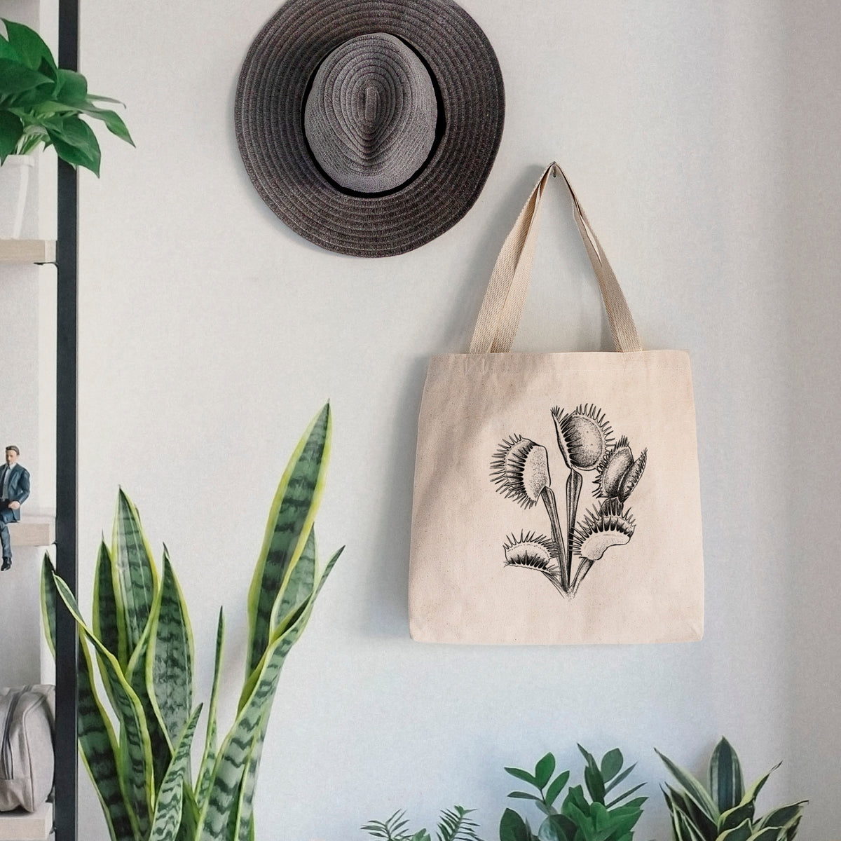 Canvas tote bag printed with a black ink illustration of a Venus flytrap plant with open traps, hanging on a wall hook below a hat, next to potted plants.