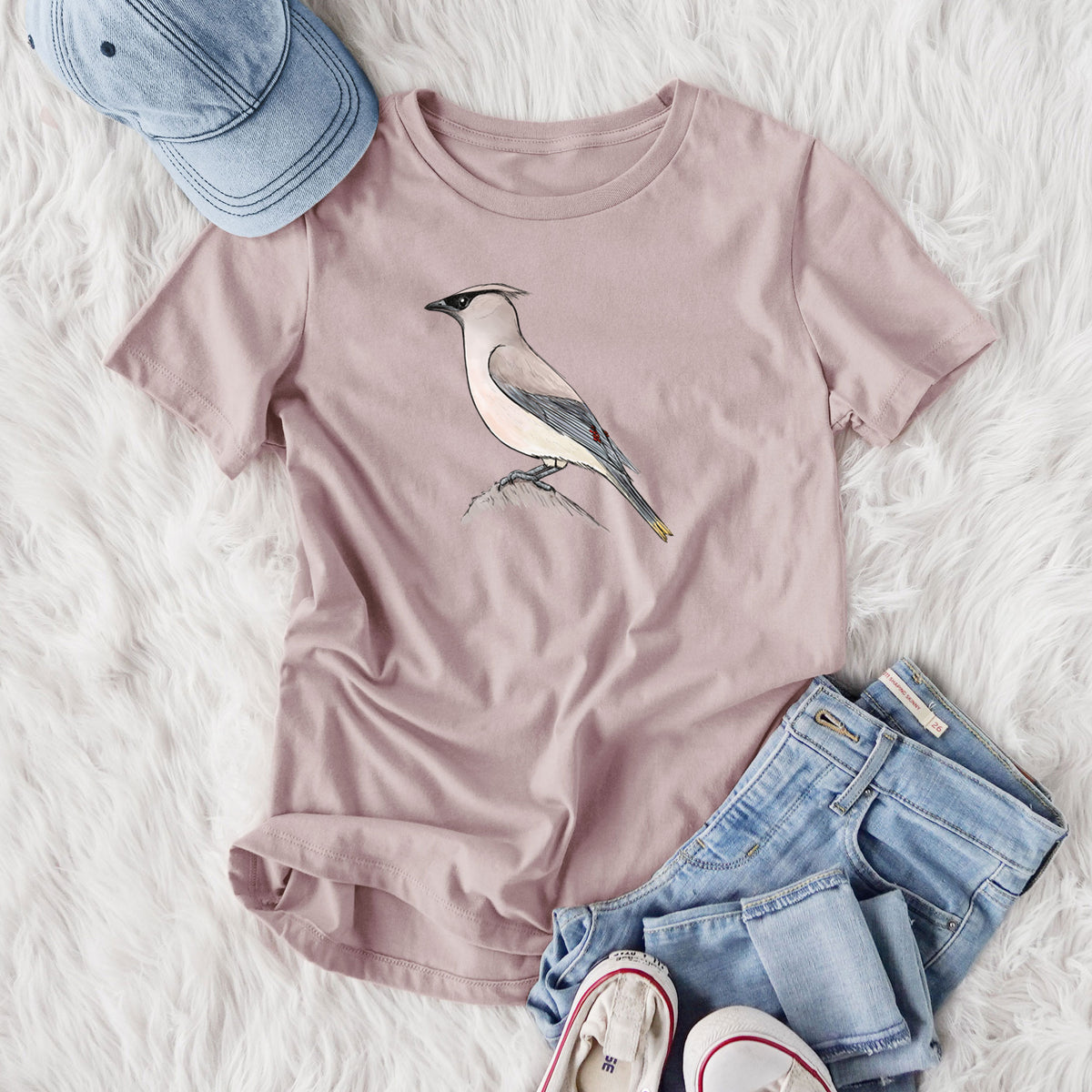 Women's relaxed fit cotton t-shirt printed with a full-color illustration of a cedar waxwing perched on a branch, styled flat on a white faux fur rug with jeans.