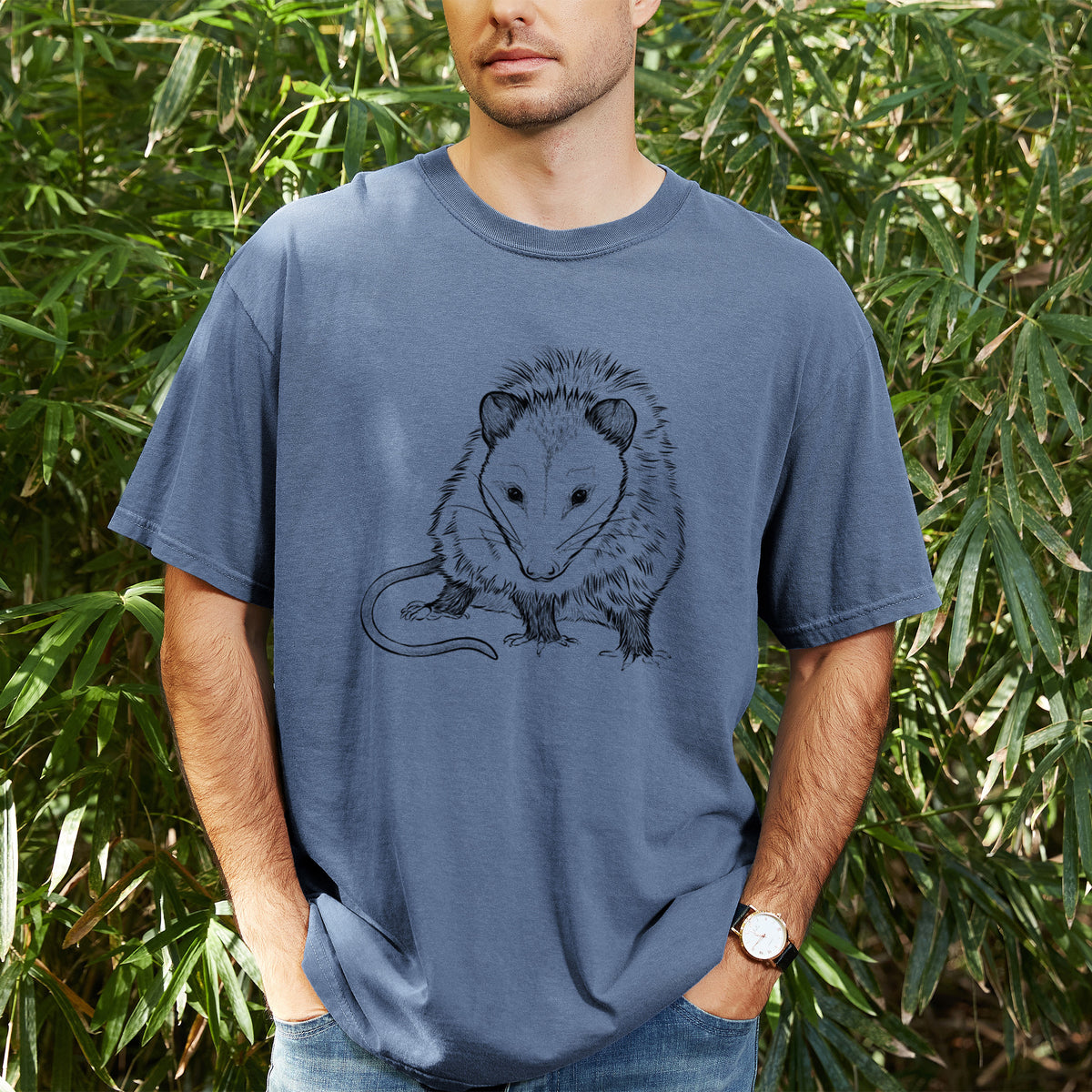 Man wearing a Comfort Colors t-shirt printed with a detailed black ink illustration of a Virginia opossum standing with its long tail curled, outdoors.