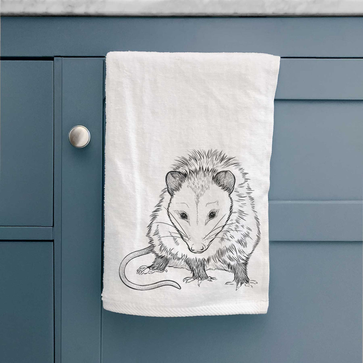 White hand towel printed with a detailed black ink illustration of a Virginia opossum standing with its long tail curled, hanging on a cabinet door.