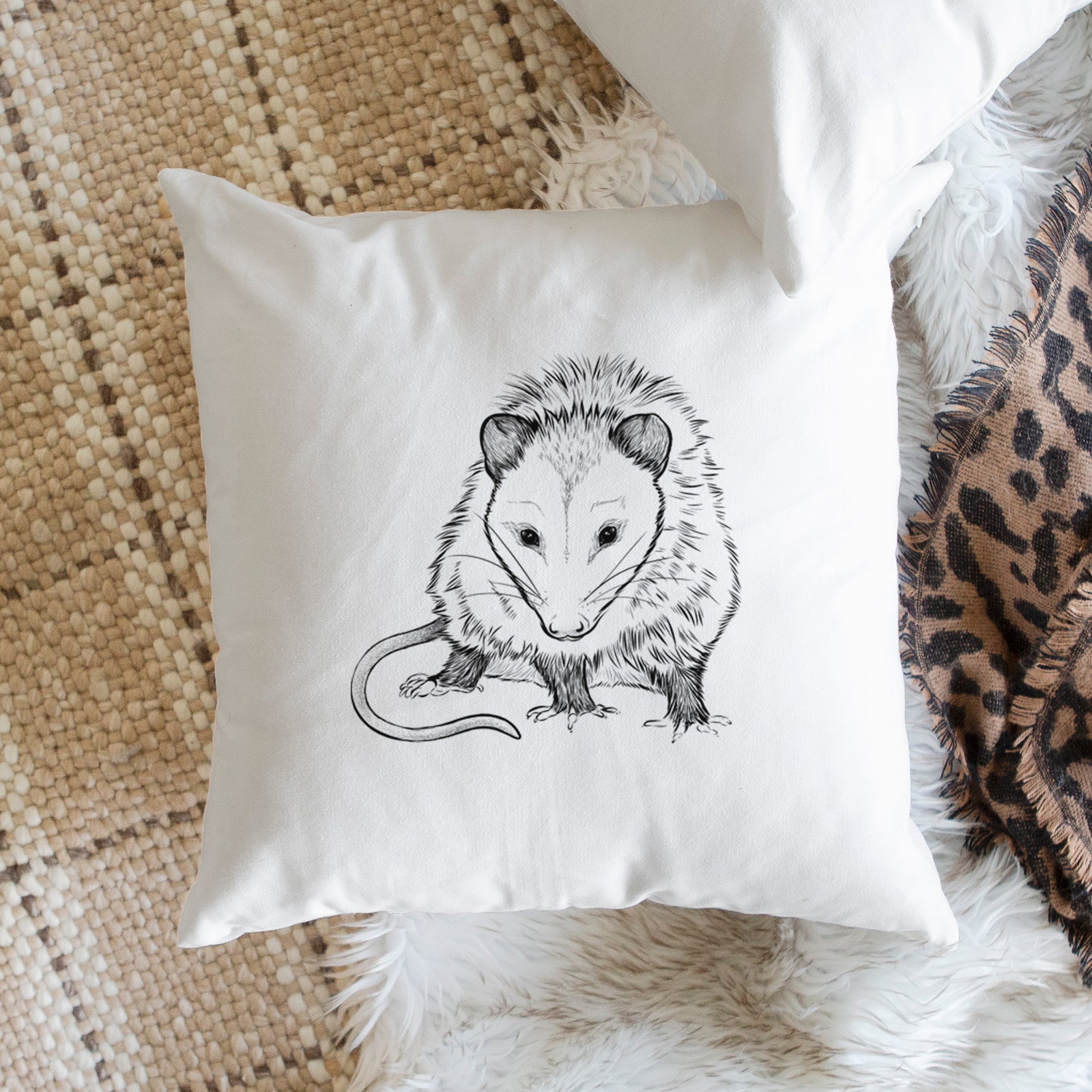 White pillow cover printed with a detailed black ink illustration of a Virginia opossum standing with its long tail curled, on a bench beside plants.