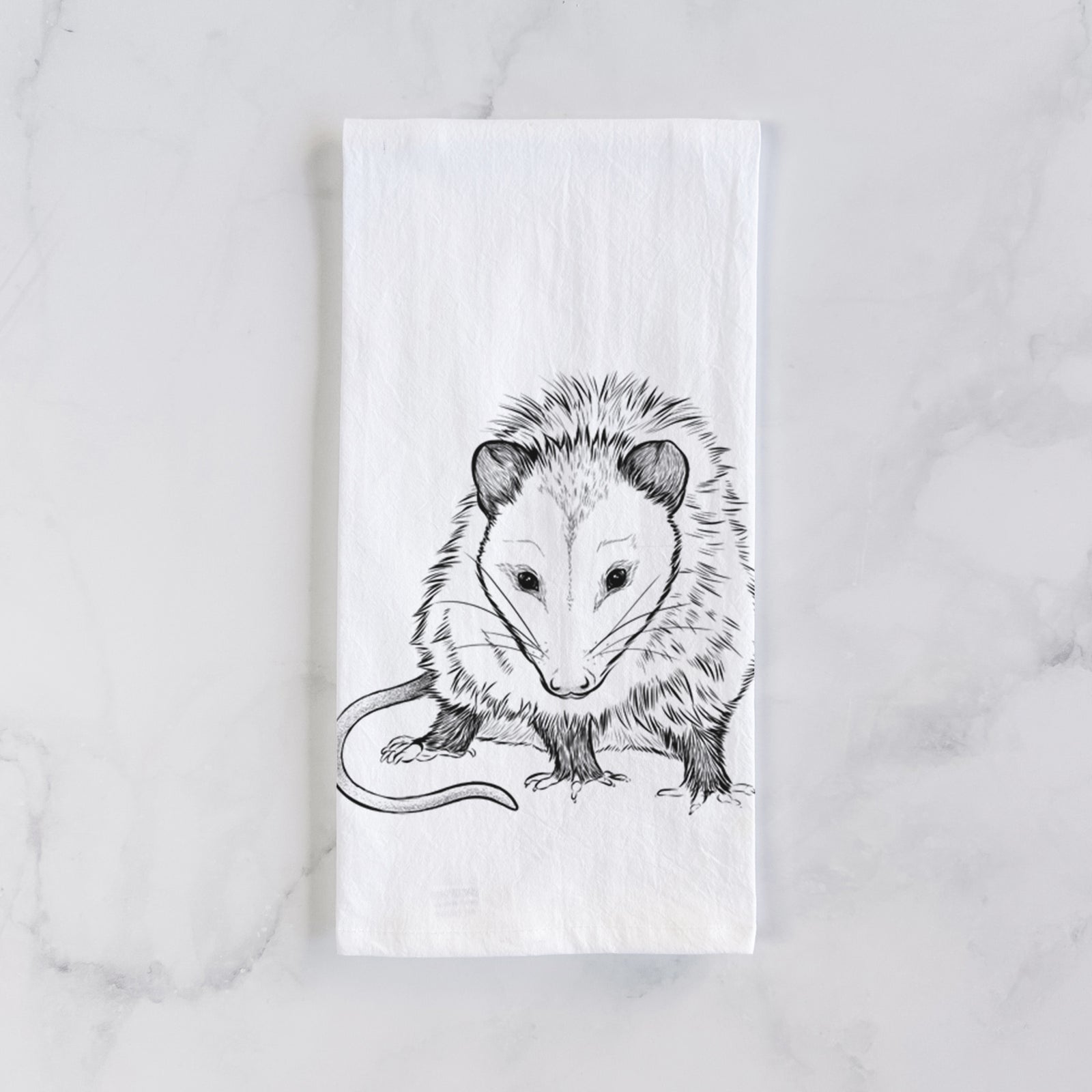 White cotton tea towel printed with a detailed black ink illustration of a Virginia opossum standing with its long tail curled, on a marble counter beside a round wooden cutting board and greenery.