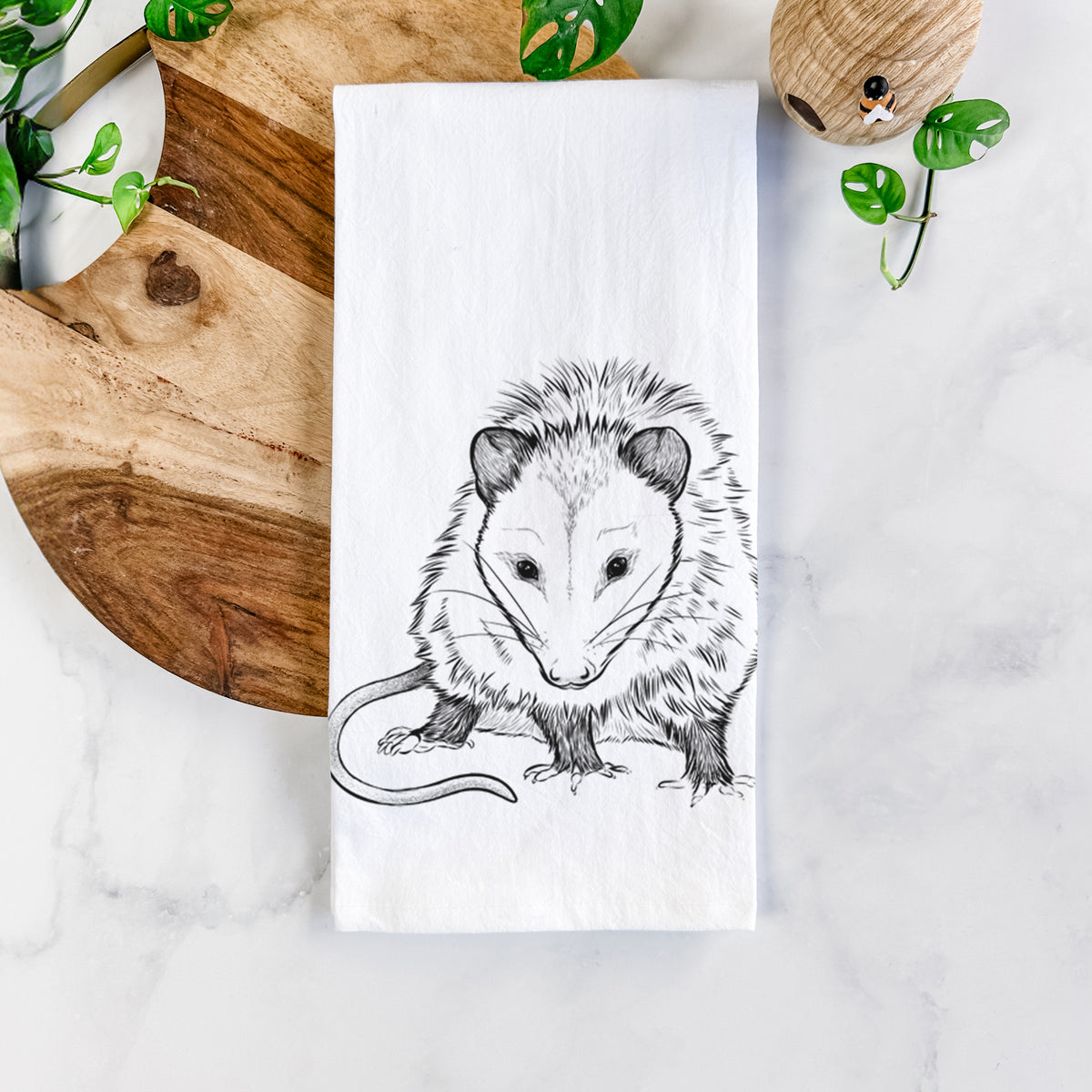 White cotton tea towel printed with a detailed black ink illustration of a Virginia opossum standing with its long tail curled, on a marble counter beside a round wooden cutting board and greenery.