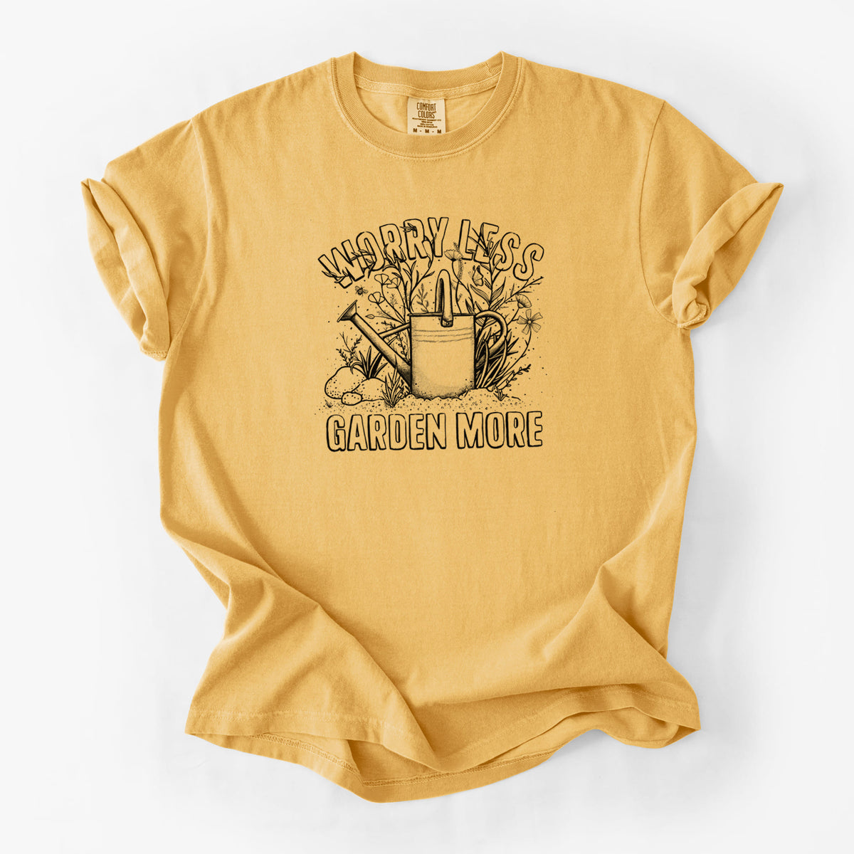 Golden wheat yellow Comfort Colors t-shirt laid flat, printed with a black ink watering can surrounded by flowers and garden tools, with the words Worry Less, Garden More.