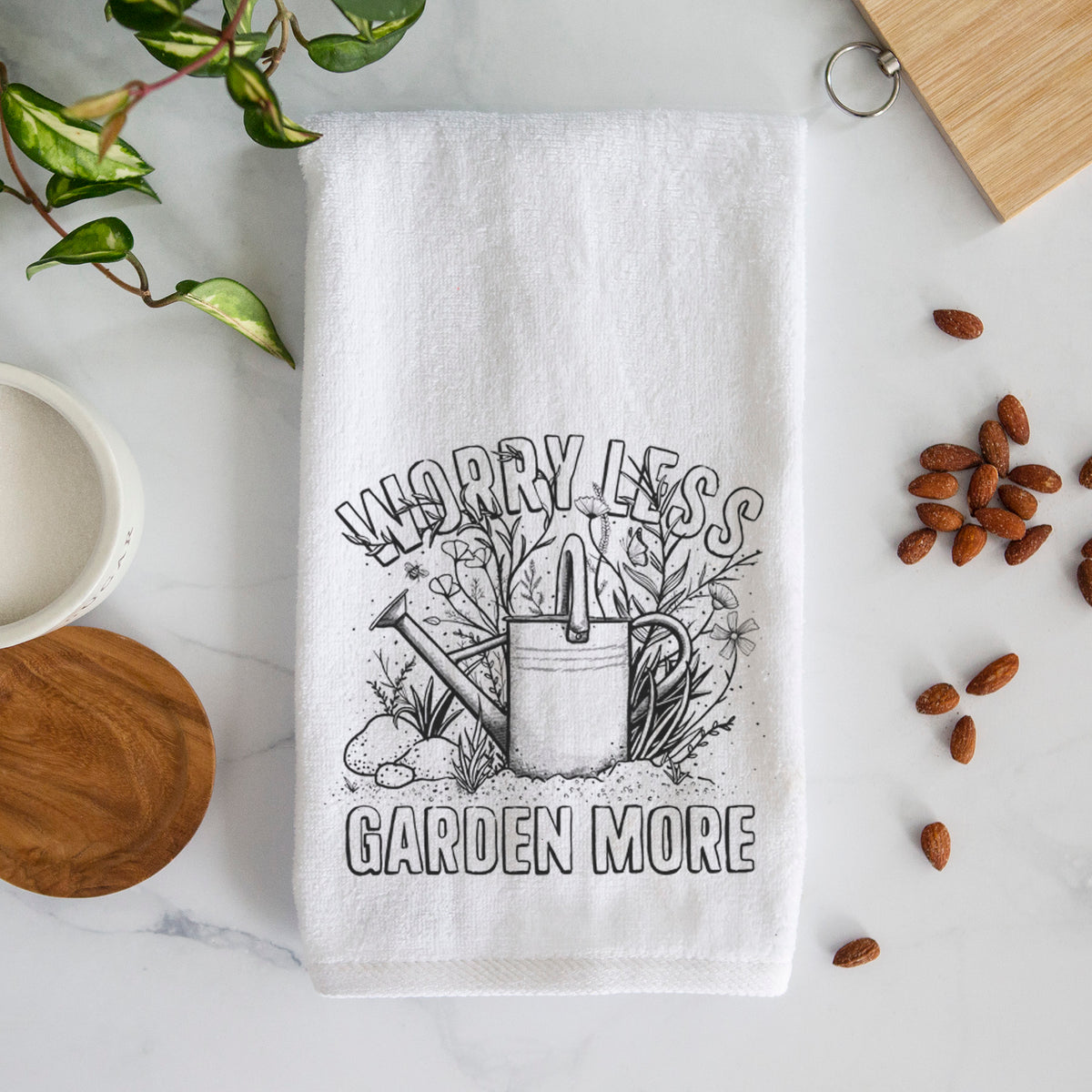 White hand towel printed with a black ink watering can surrounded by flowers and garden tools, with the words Worry Less, Garden More.