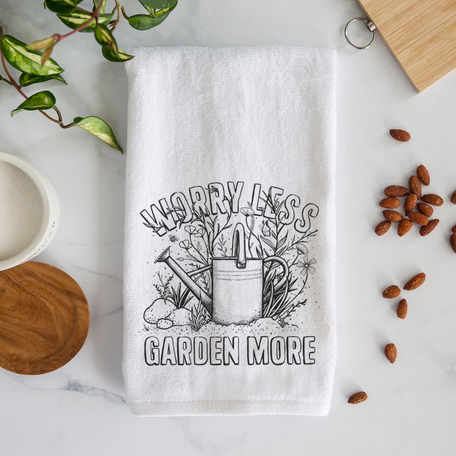 White hand towel printed with a black ink watering can surrounded by flowers and garden tools, with the words Worry Less, Garden More.