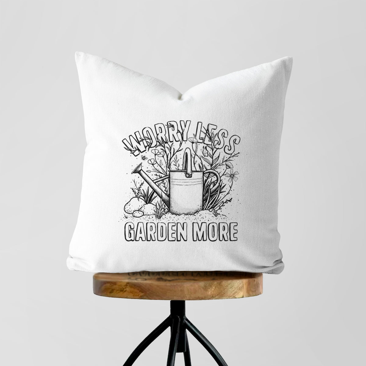 White pillow cover printed with a black ink watering can surrounded by flowers and garden tools, with the words Worry Less, Garden More, on a wooden stool.