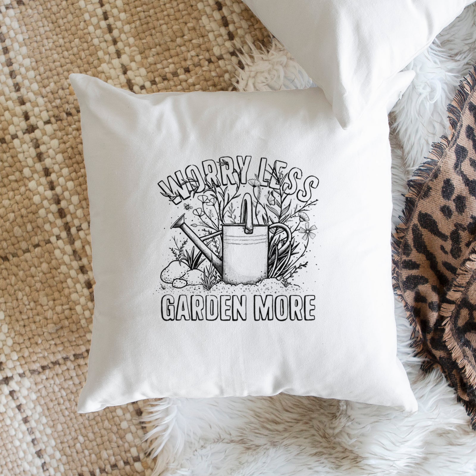 White pillow cover printed with a black ink watering can surrounded by flowers and garden tools, with the words Worry Less, Garden More, on a bench beside plants.