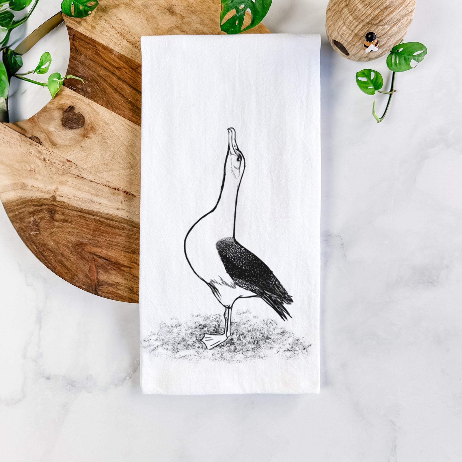 White cotton tea towel printed with a black ink illustration of a wandering albatross with its head raised, on a marble counter beside a round wooden cutting board and greenery.