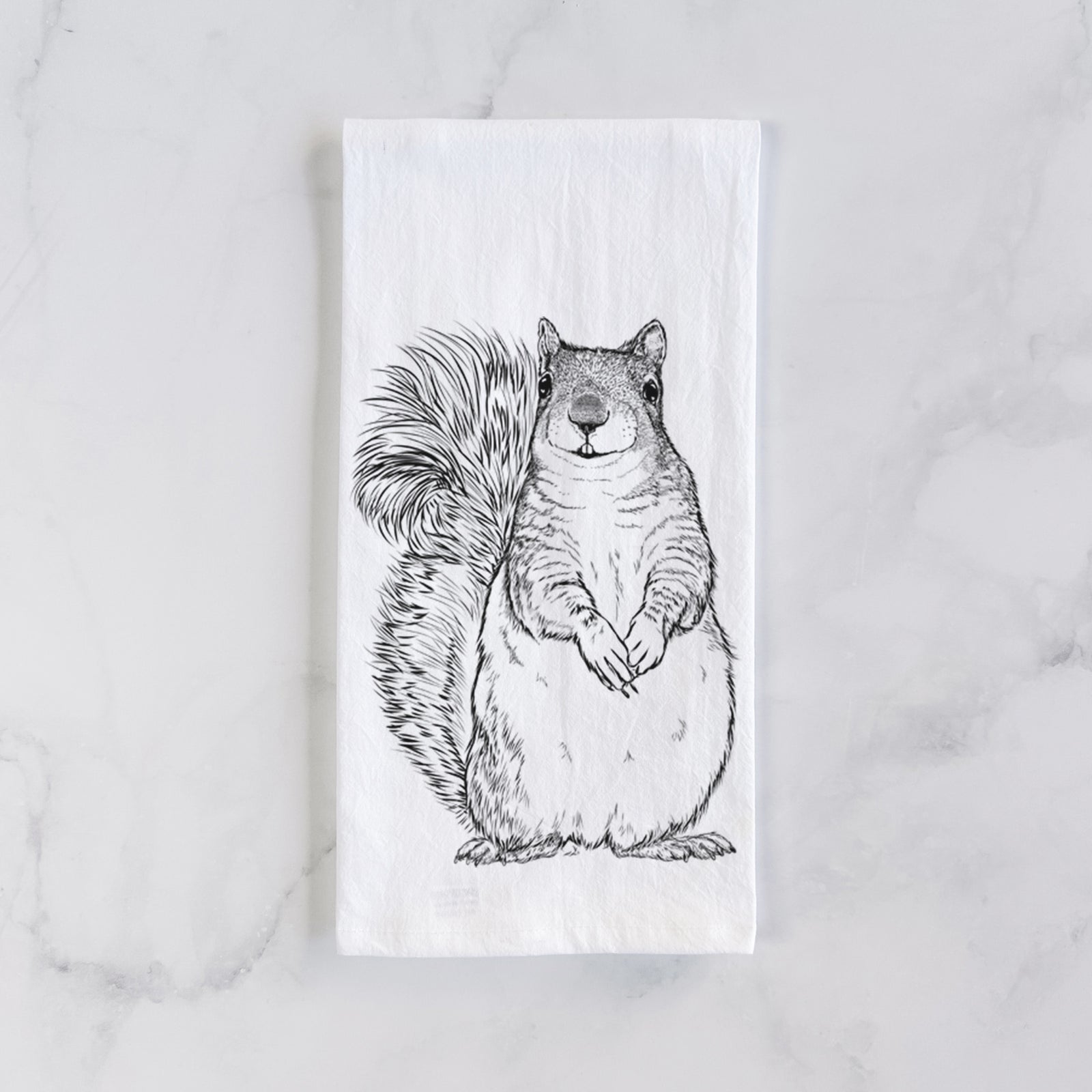 White cotton tea towel printed with a detailed black ink illustration of a western grey squirrel sitting upright with a bushy tail, on a marble counter beside a round wooden cutting board and greenery.