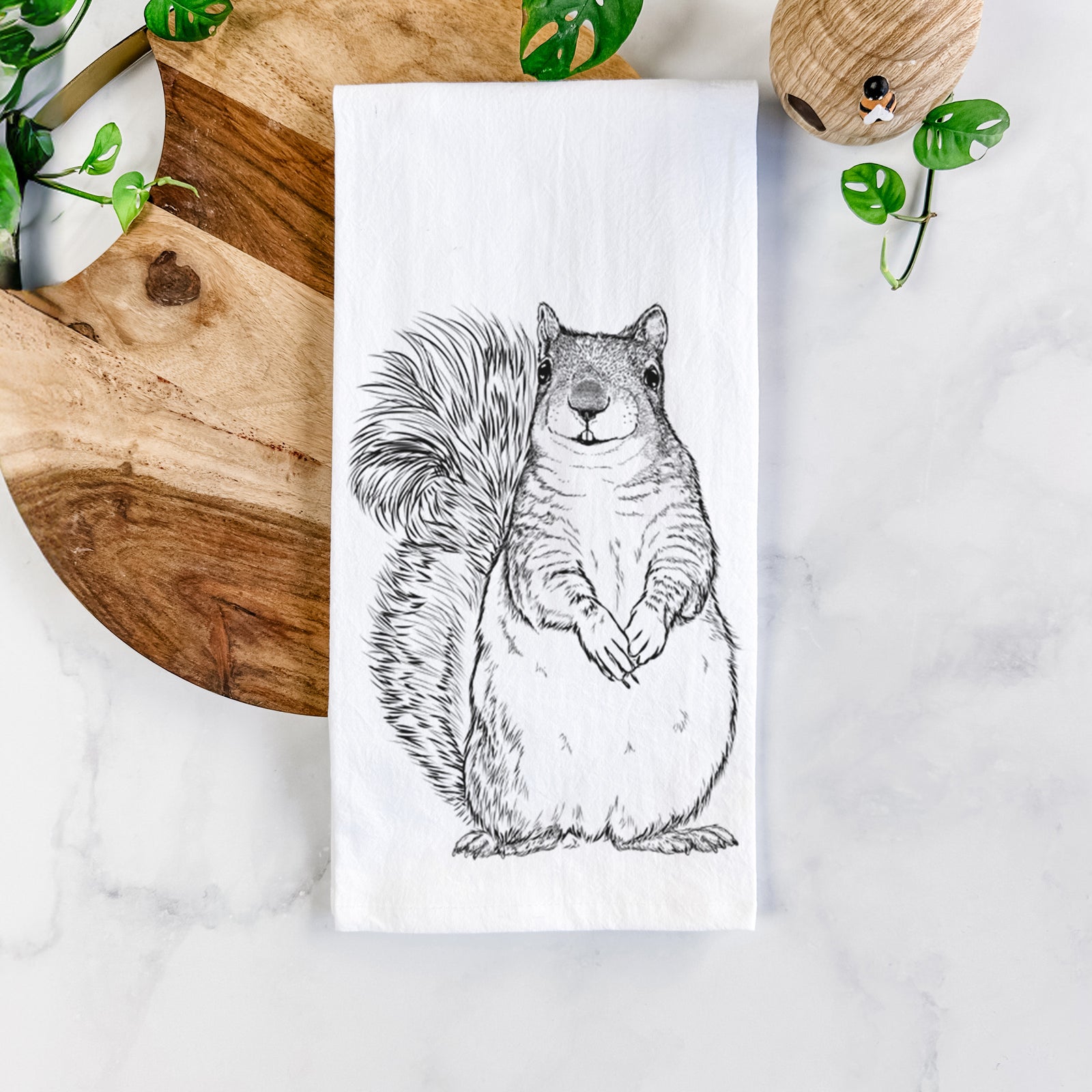 White cotton tea towel printed with a detailed black ink illustration of a western grey squirrel sitting upright with a bushy tail, on a marble counter beside a round wooden cutting board and greenery.
