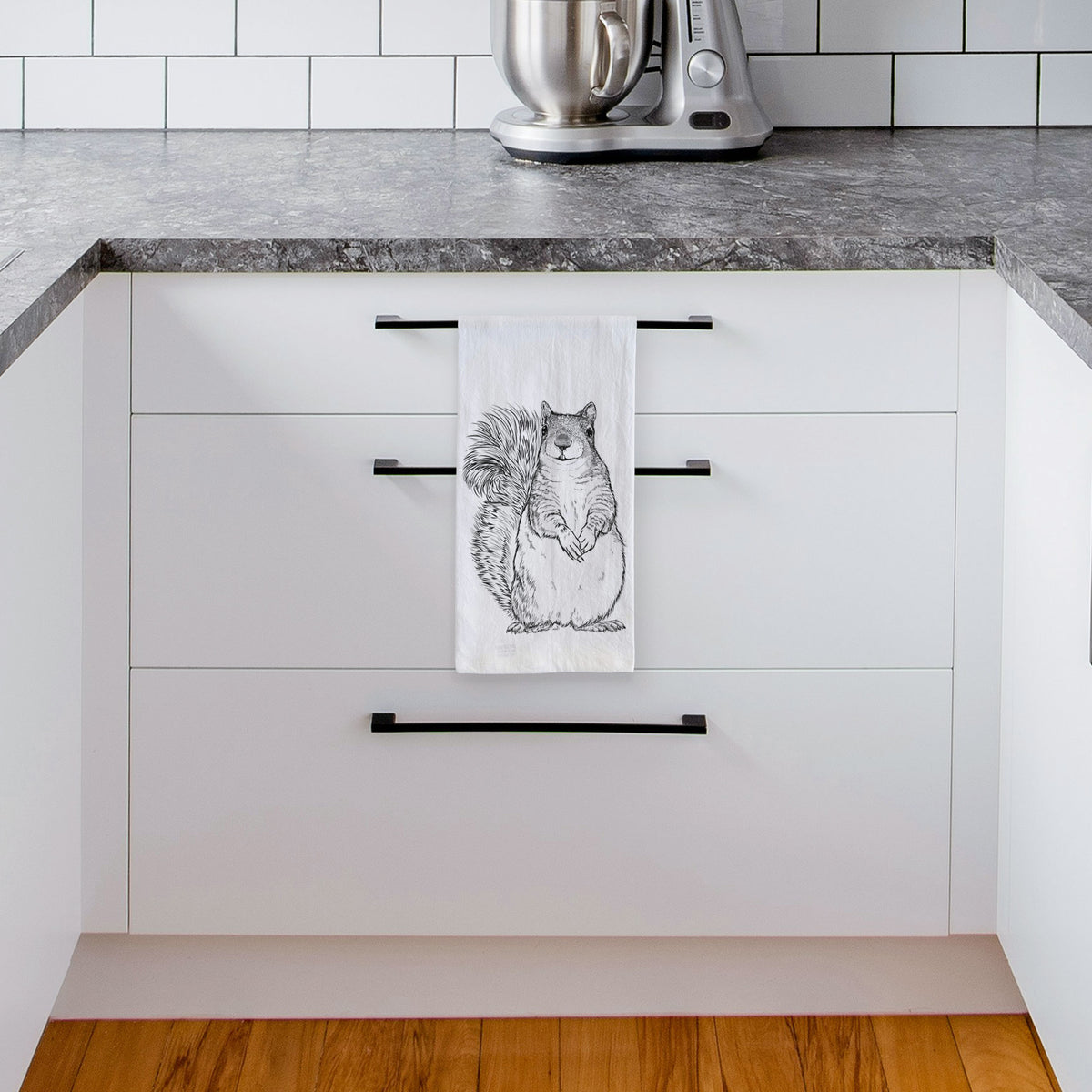 White tea towel printed with a detailed black ink illustration of a western grey squirrel sitting upright with a bushy tail, hanging from a drawer handle in a bright white kitchen.