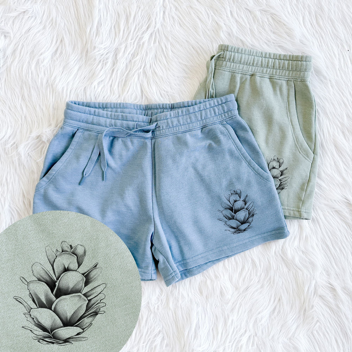 Two pairs of Cali Wave shorts in different colors printed with a detailed black ink illustration of a western hemlock pine cone, with a close-up of the print.