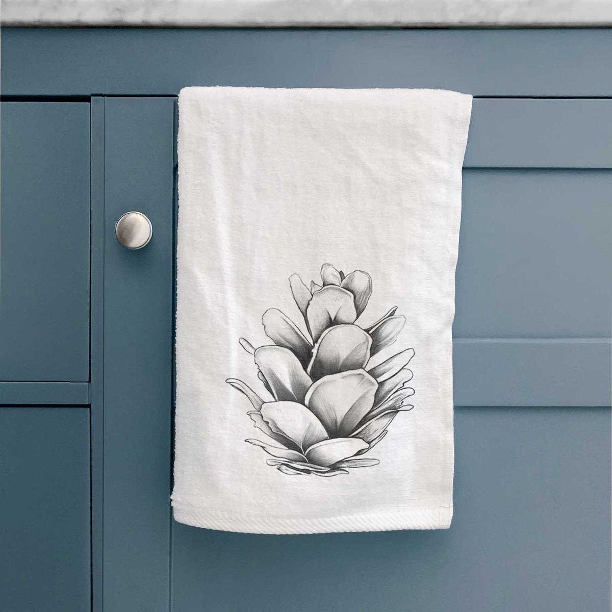 White hand towel printed with a detailed black ink illustration of a western hemlock pine cone, hanging on a cabinet door.