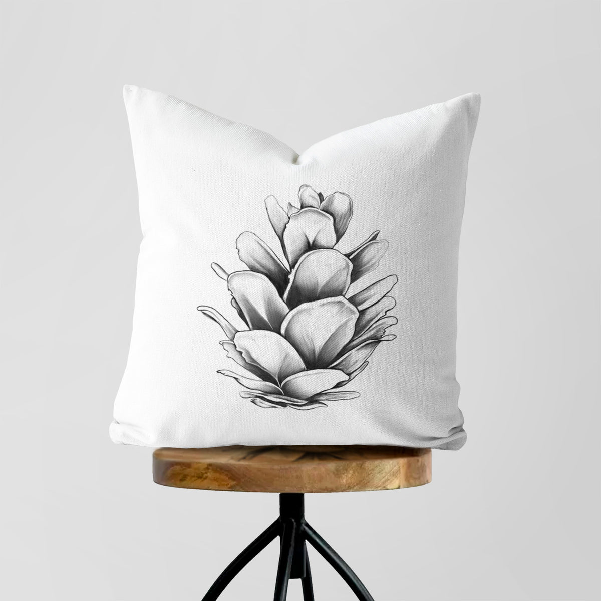 White pillow cover printed with a detailed black ink illustration of a western hemlock pine cone, on a wooden stool.