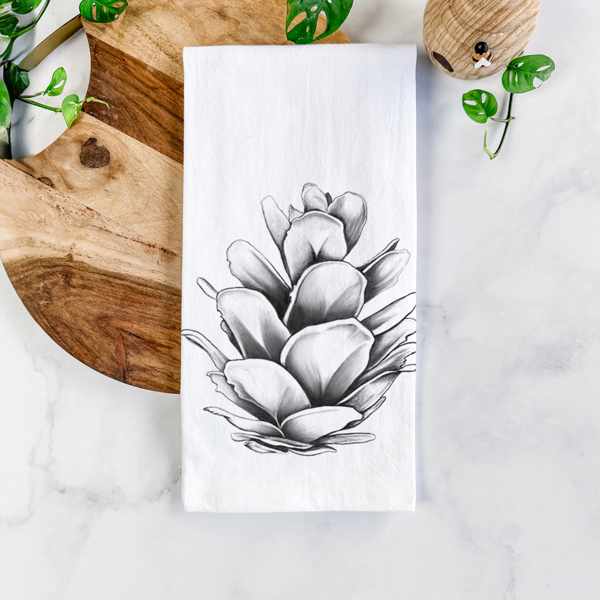 White cotton tea towel printed with a detailed black ink illustration of a western hemlock pine cone, on a marble counter beside a round wooden cutting board and greenery.