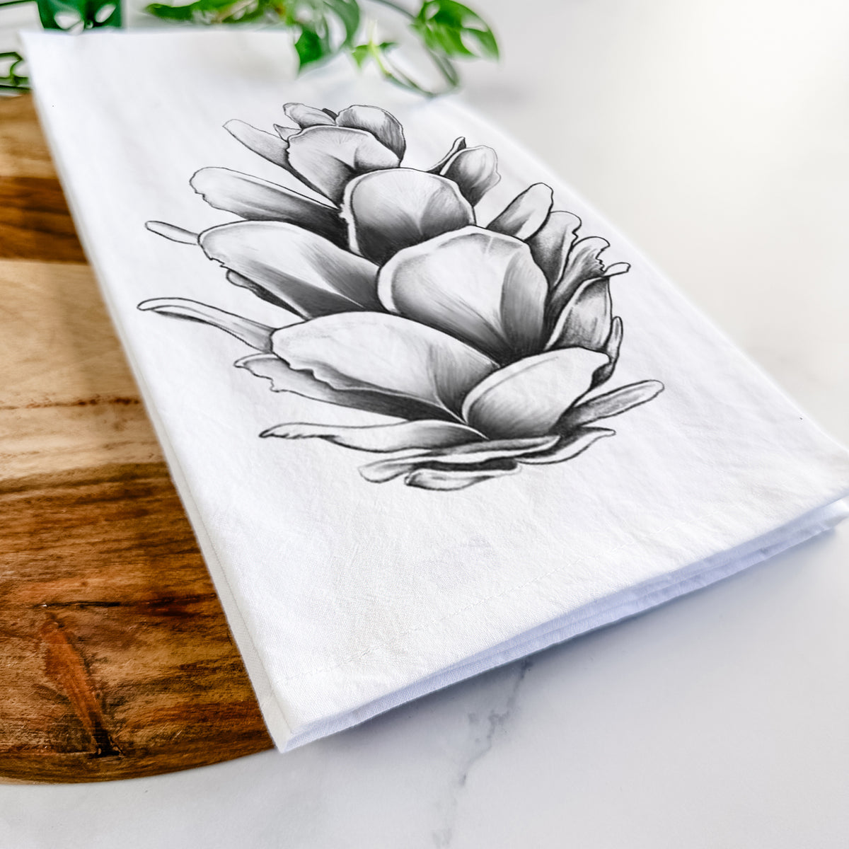 Close-up of a folded white tea towel on a wooden board, showing a detailed black ink illustration of a western hemlock pine cone.