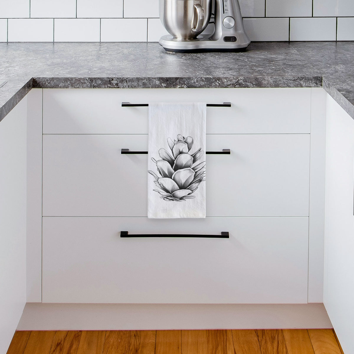 White tea towel printed with a detailed black ink illustration of a western hemlock pine cone, hanging from a drawer handle in a bright white kitchen.