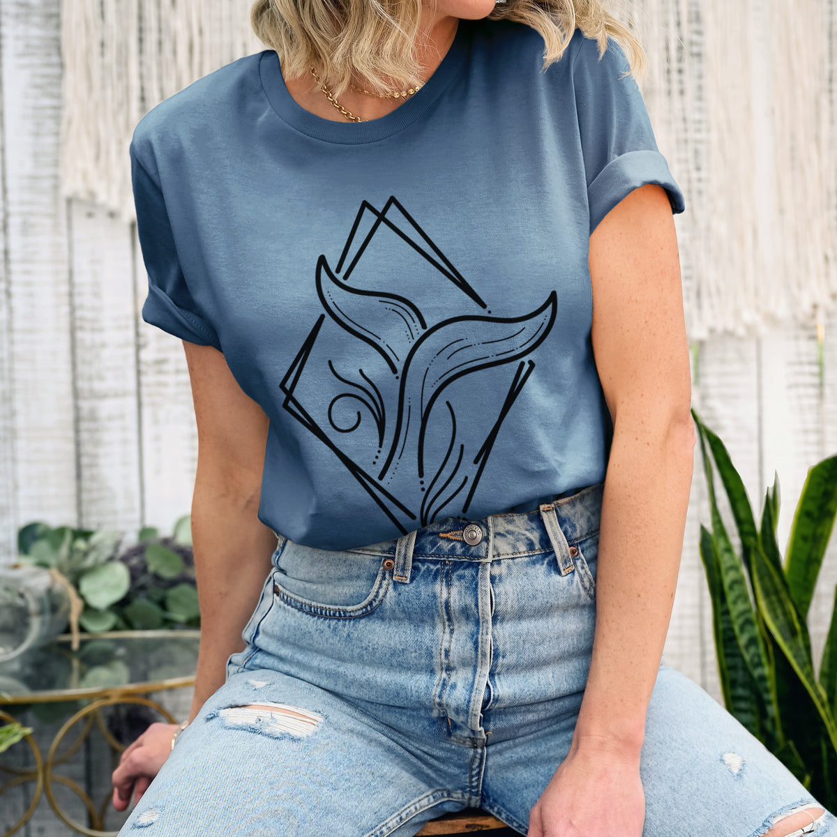 Woman wearing a steel blue lightweight cotton t-shirt printed with a black line art whale tail rising from a wave inside a diamond outline.