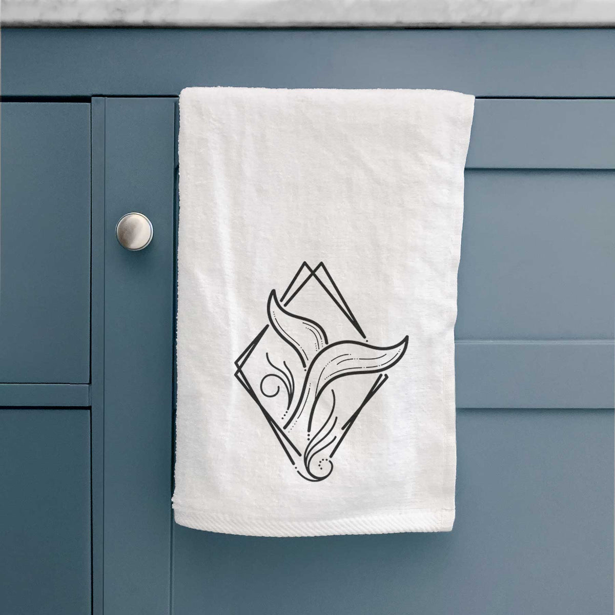 White hand towel printed with a black line art whale tail rising from a wave inside a diamond outline, hanging on a cabinet door.