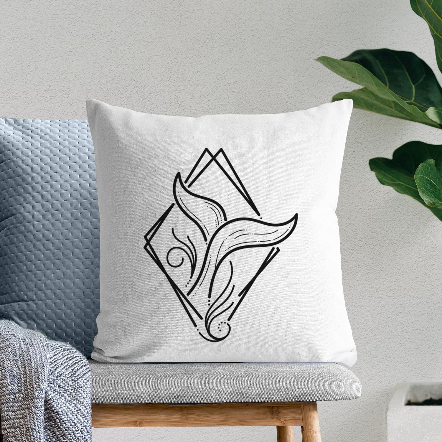 White pillow cover printed with a black line art whale tail rising from a wave inside a diamond outline, on a bench beside plants.
