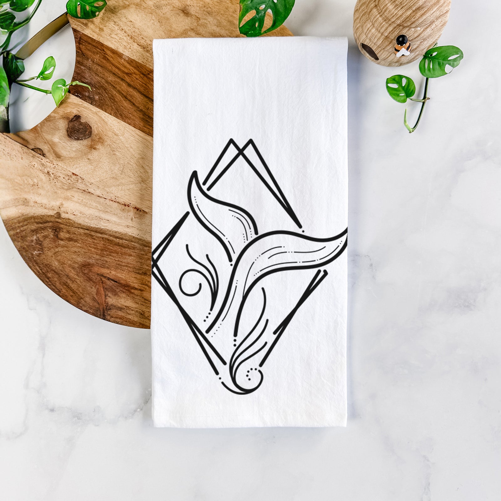 White cotton tea towel printed with a black line art whale tail rising from a wave inside a diamond outline, on a marble counter beside a round wooden cutting board and greenery.