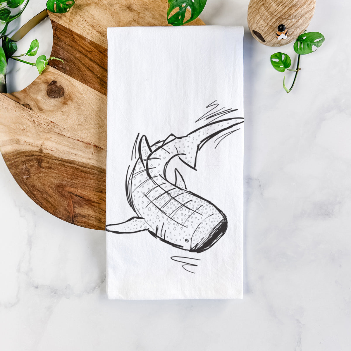 White cotton tea towel printed with a black ink illustration of a spotted whale shark swimming, on a marble counter beside a round wooden cutting board and greenery.