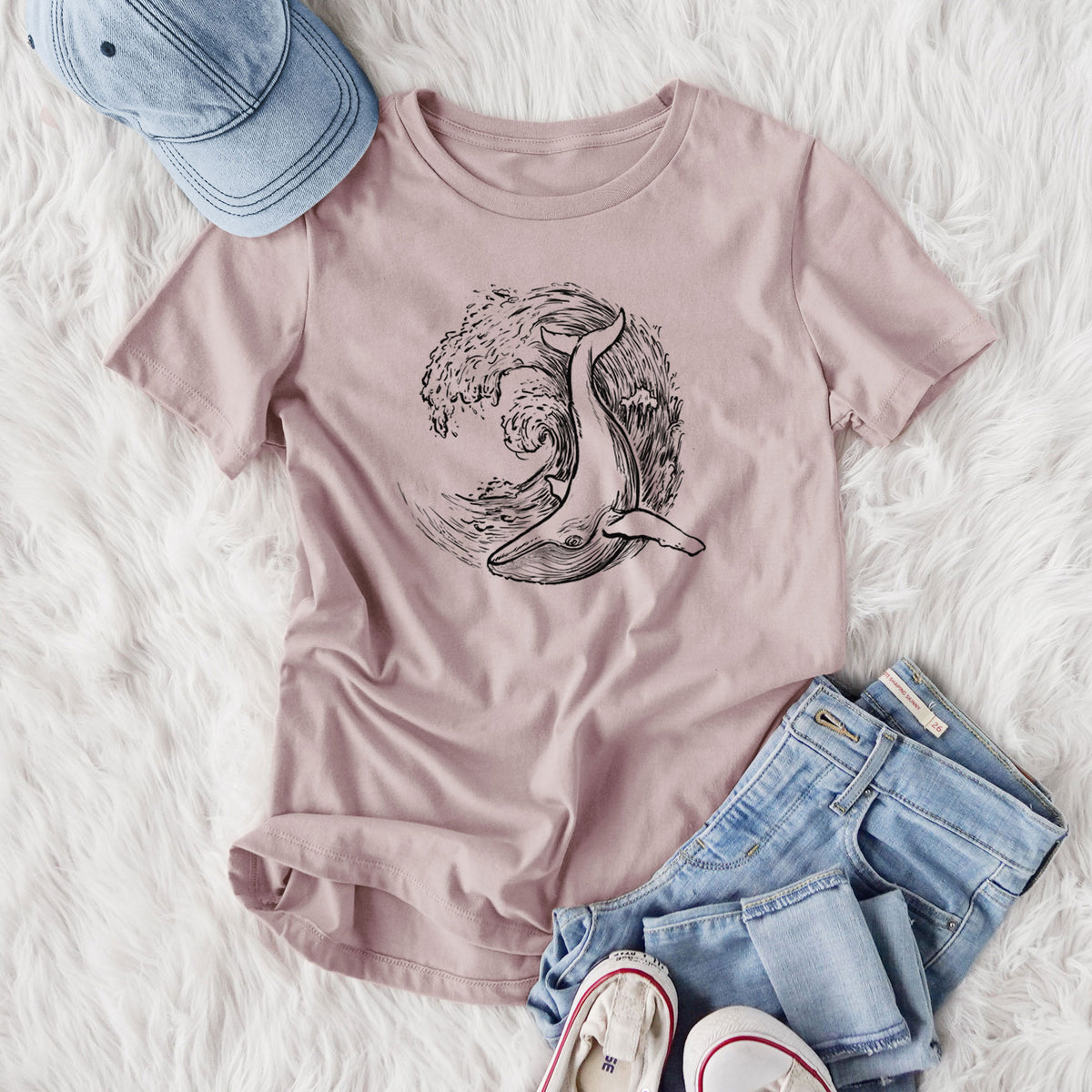 Women's relaxed fit cotton t-shirt printed with a black ink illustration of a humpback whale swimming inside a curling ocean wave, styled flat on a white faux fur rug with jeans.