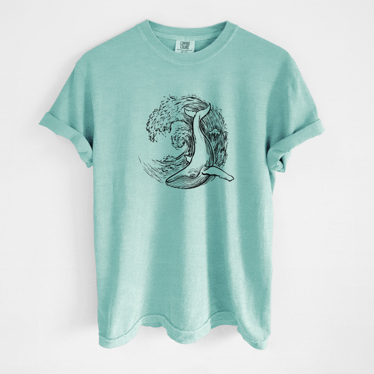 Chalky mint Comfort Colors t-shirt laid flat, printed with a black ink illustration of a humpback whale swimming inside a curling ocean wave.