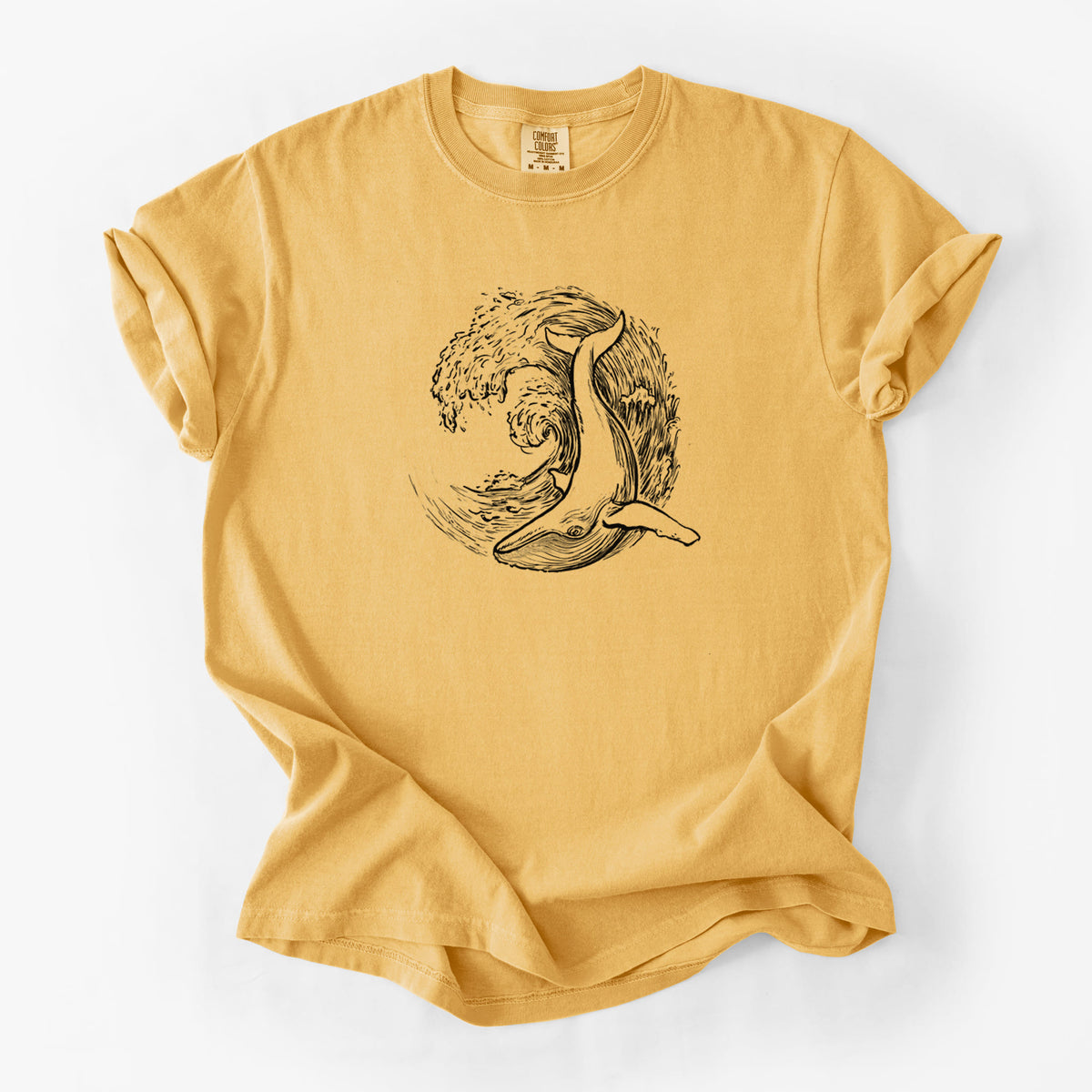 Golden wheat yellow Comfort Colors t-shirt laid flat, printed with a black ink illustration of a humpback whale swimming inside a curling ocean wave.