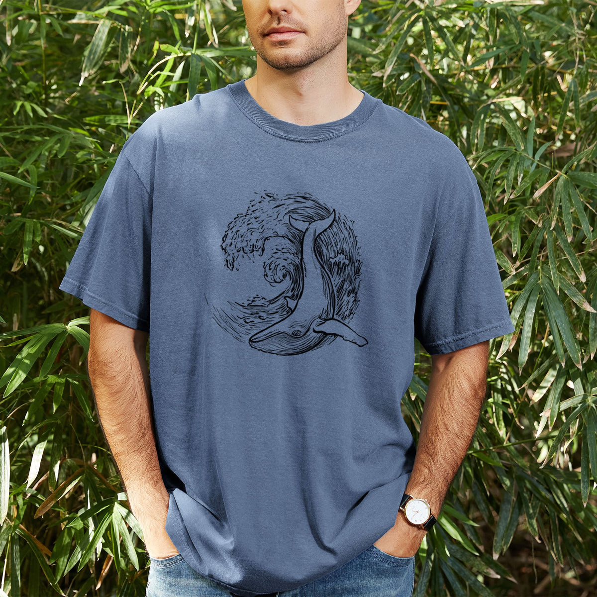 Man wearing a Comfort Colors t-shirt printed with a black ink illustration of a humpback whale swimming inside a curling ocean wave, outdoors.