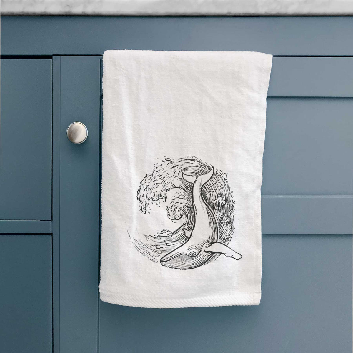 White hand towel printed with a black ink illustration of a humpback whale swimming inside a curling ocean wave, hanging on a cabinet door.
