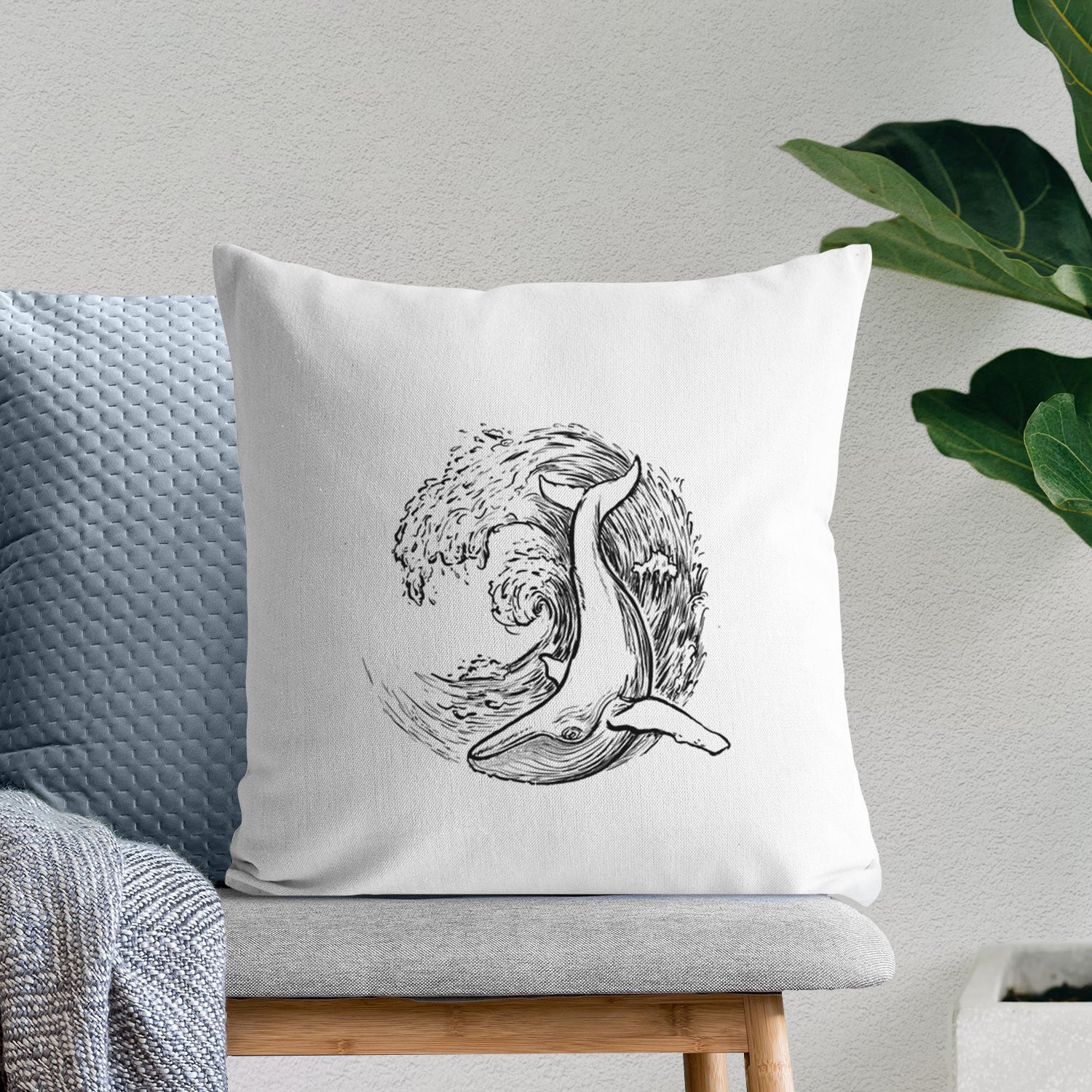 White pillow cover printed with a black ink illustration of a humpback whale swimming inside a curling ocean wave, on a bench beside plants.