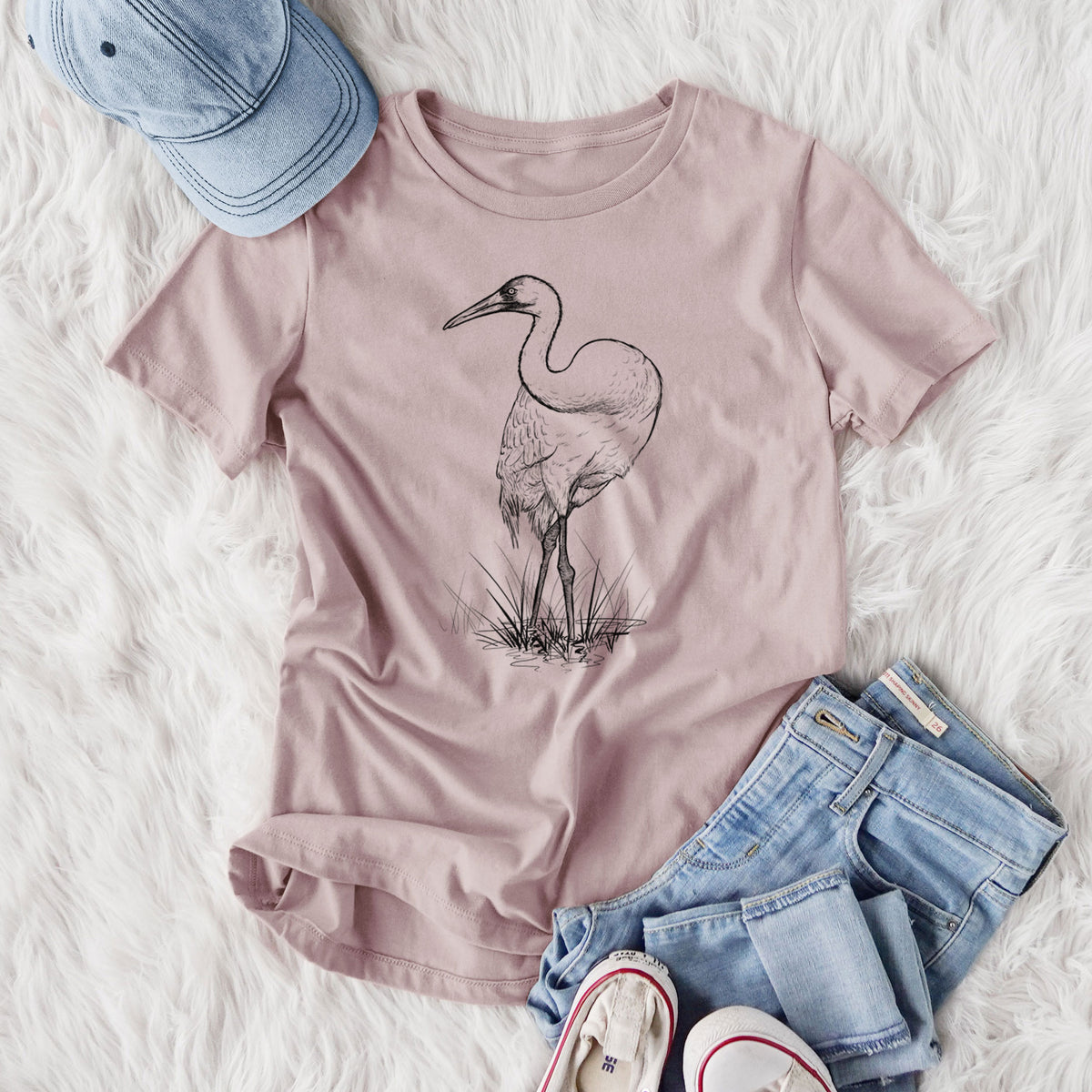Women's relaxed fit cotton t-shirt printed with a detailed black ink illustration of a whooping crane standing in grass, styled flat on a white faux fur rug with jeans.