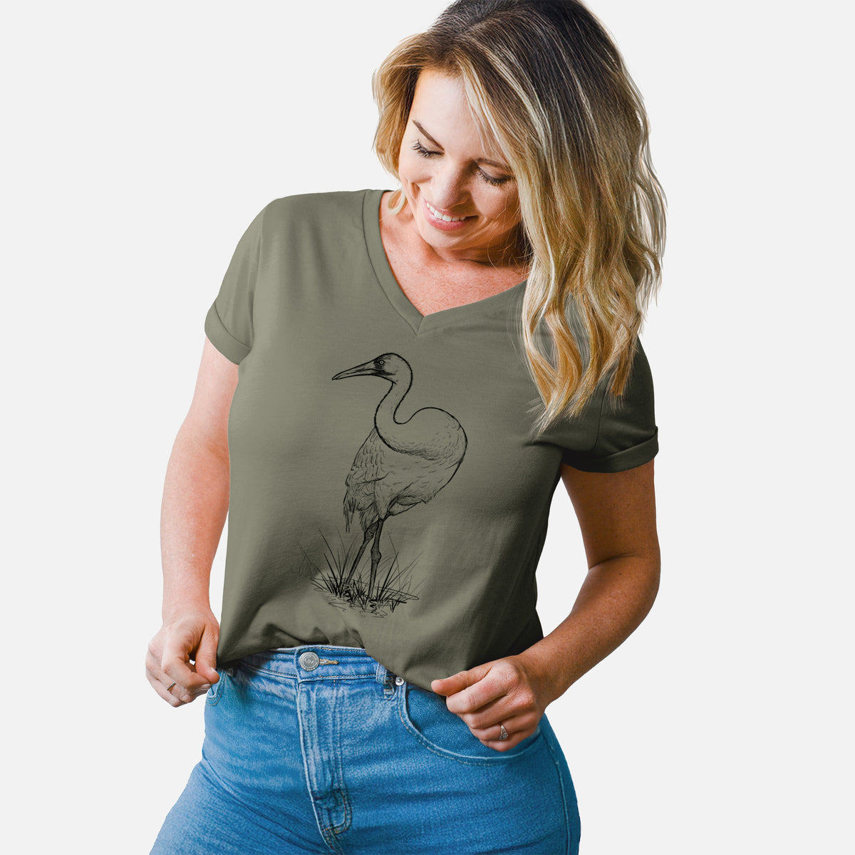 Woman wearing a military green women's relaxed fit cotton V-neck t-shirt printed with a detailed black ink illustration of a whooping crane standing in grass.