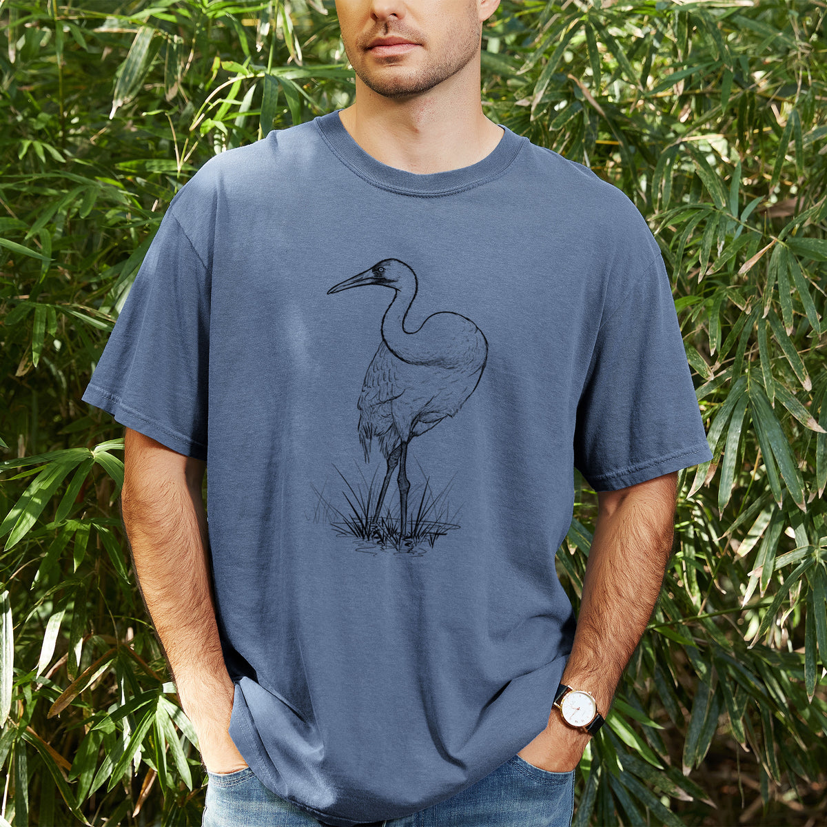 Man wearing a Comfort Colors t-shirt printed with a detailed black ink illustration of a whooping crane standing in grass, outdoors.