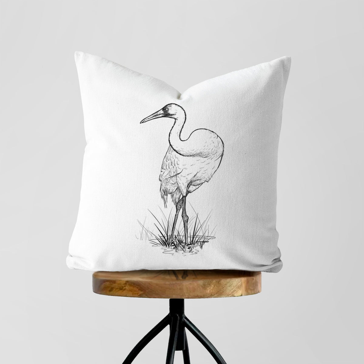 White pillow cover printed with a detailed black ink illustration of a whooping crane standing in grass, on a wooden stool.