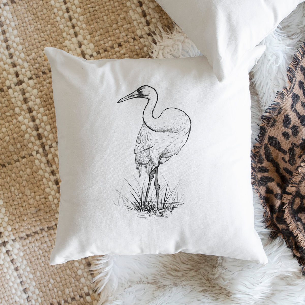 White pillow cover printed with a detailed black ink illustration of a whooping crane standing in grass, on a woven chair.
