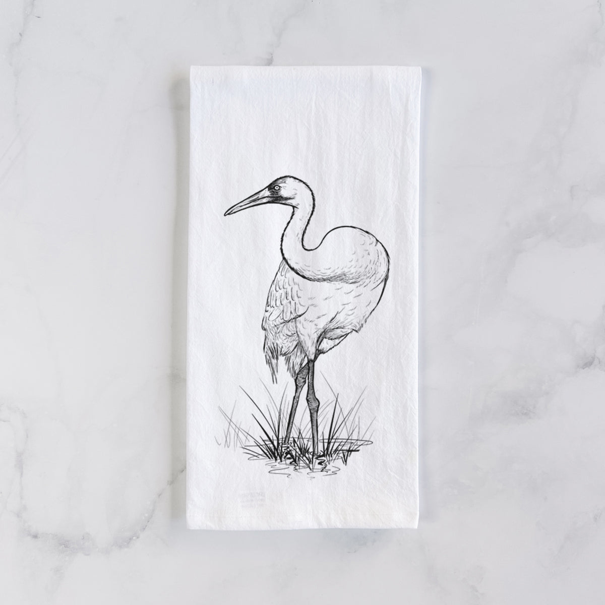 White cotton tea towel laid flat on marble, printed with a detailed black ink illustration of a whooping crane standing in grass.