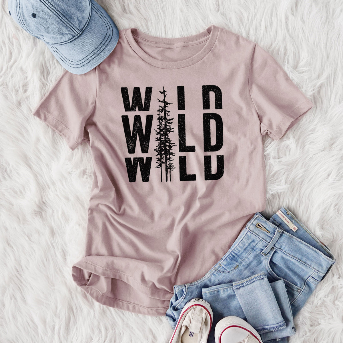 Women's relaxed fit cotton t-shirt printed with bold black text reading WILD stacked three times, with a tall pine tree forming the letter I, styled flat on a white faux fur rug with jeans.